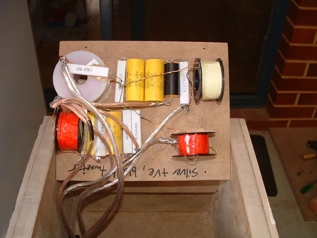 DIY speaker crossover wired point-to-point on an MDF board: air-core inductors, capacitors, and power resistors with speaker wire, handwritten tweeter polarity note, board resting on an open cabinet.