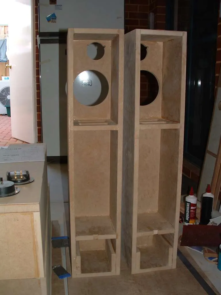 Two unfinished MDF floorstanding speaker cabinets side by side in a workshop, internal bracing and tweeter and woofer cutouts visible before front baffles are fitted.