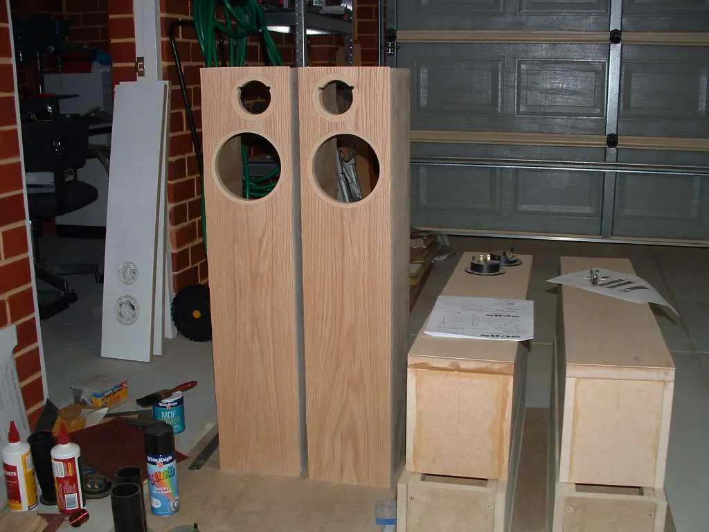 Speaker build in a garage: two veneered floorstanders with driver cutouts standing upright, two raw MDF cabinets on their sides, wood glue, MDF primer, and port tubes nearby.